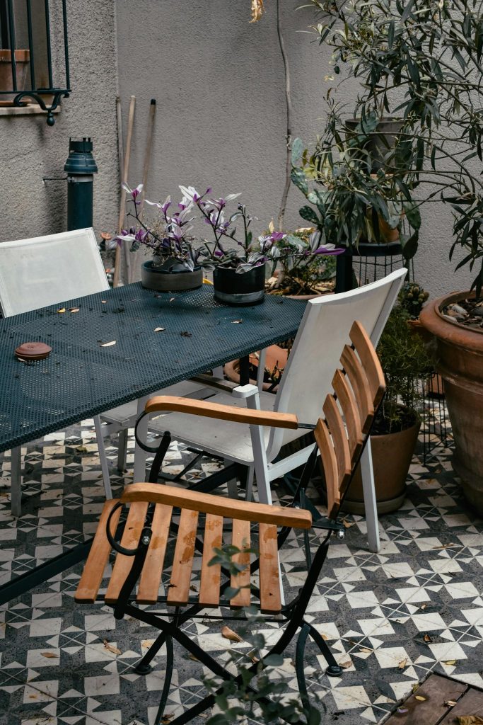 Rustic patio setting with wooden chairs, a table, and lush potted plants.